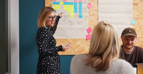 Woman Pointing at Post-it Notes on Wall While Female Colleague Looks On